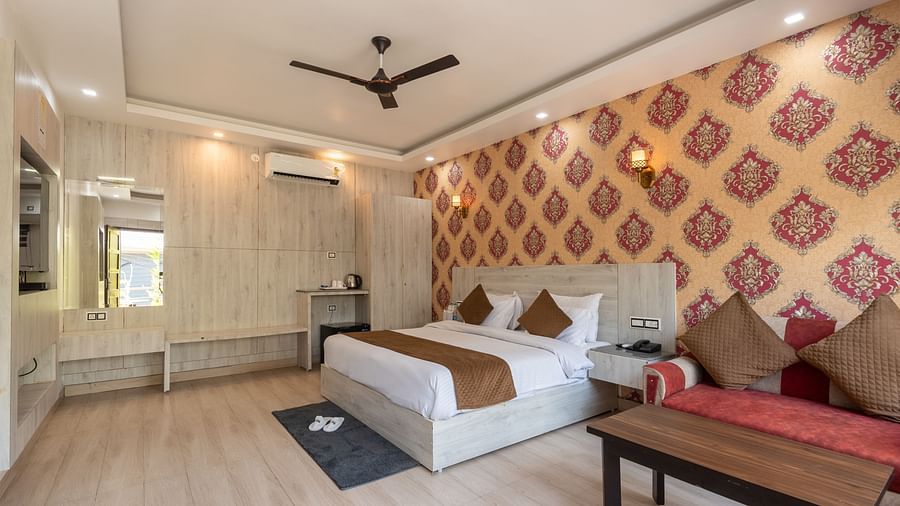 Spacious, well-lit hotel room with a large double bed, patterned feature wall in red and gold, modern furnishings, ceiling fan, and a comfortable seating area with a coffee table - DLS Nature Trinket Resort, Dalhousie.