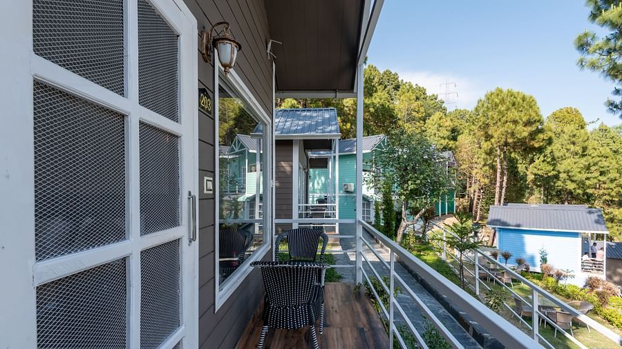 Private balcony with outdoor seating overlooking a peaceful resort area, surrounded by colourful cottages and tall pine trees under a clear blue sky - DLS Nature Trinket Resort, Dalhousie.