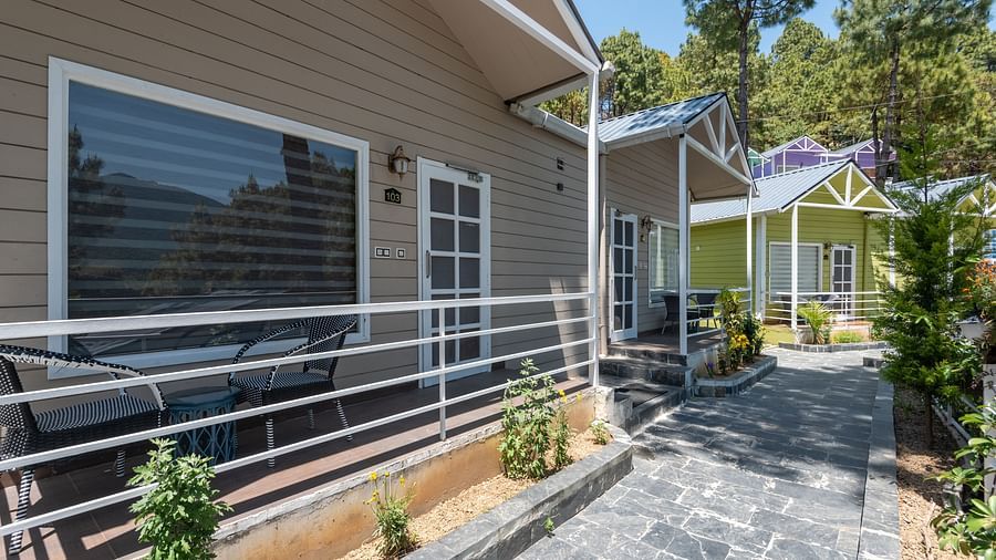  Charming row of modern wooden cottages with white railings and patio seating, set along a paved walkway surrounded by pine trees and landscaped greenery - DLS Nature Trinket Resort, Dalhousie.
