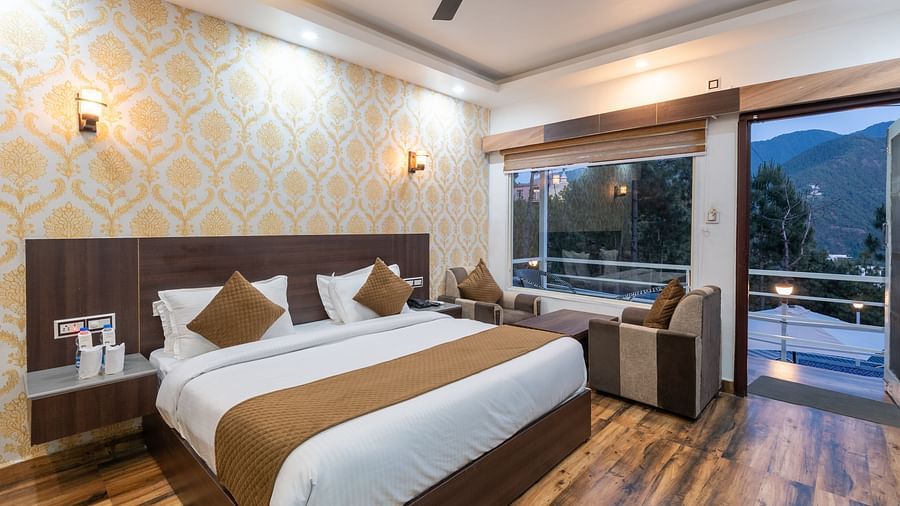 Elegant hotel room with a large double bed, warm wooden flooring, patterned wallpaper, a sitting area by a large window, and a balcony offering scenic views of the mountains and trees - DLS Nature Trinket Resort, Dalhousie.