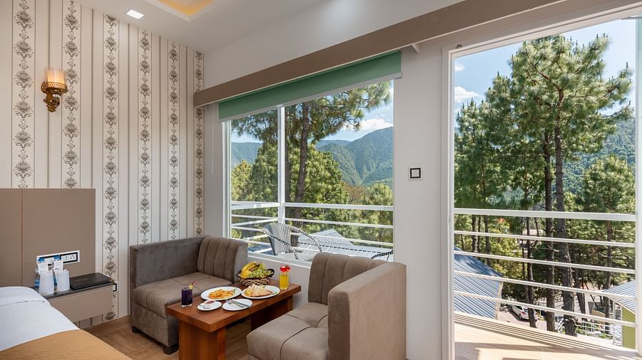 Bright and modern hotel room with a small seating area, breakfast set on a coffee table, and large windows showcasing a scenic mountain and pine forest view with access to a balcony - DLS Nature Trinket Resort, Dalhousie.