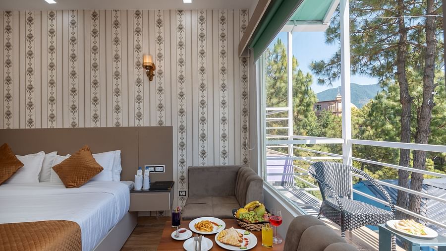 Comfortable hotel room featuring a neatly made bed, a sitting area with food and drinks on a coffee table, and a large window with a view of pine trees and distant mountains from the balcony - DLS Nature Trinket Resort, Dalhousie.
