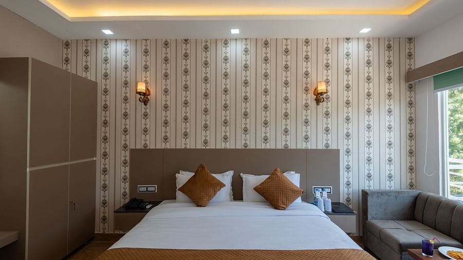 Elegant hotel bedroom with a double bed, decorative wallpaper featuring subtle floral patterns, warm lighting from wall sconces, a modern wardrobe, a side table, and a comfortable seating area by a window - DLS Nature Trinket Resort, Dalhousie.