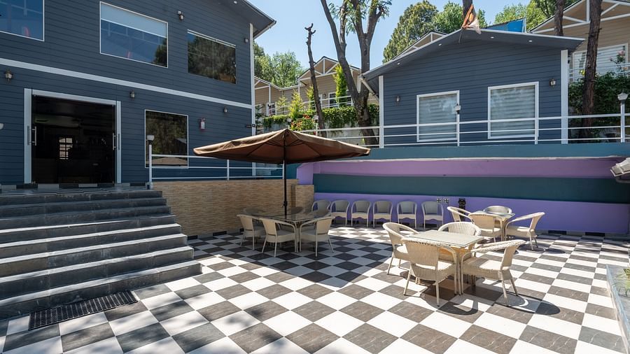Open courtyard with black-and-white checkered flooring, beige patio furniture, and a brown umbrella, surrounded by modern grey buildings and trees - DLS Nature Trinket Resort, Dalhousie.