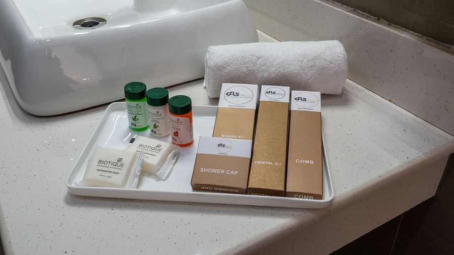 Close-up of a bathroom countertop displaying a neatly arranged tray with toiletries, including bottles, soap, and boxed items, alongside a rolled white towel - DLS Nature Trinket Resort, Dalhousie.