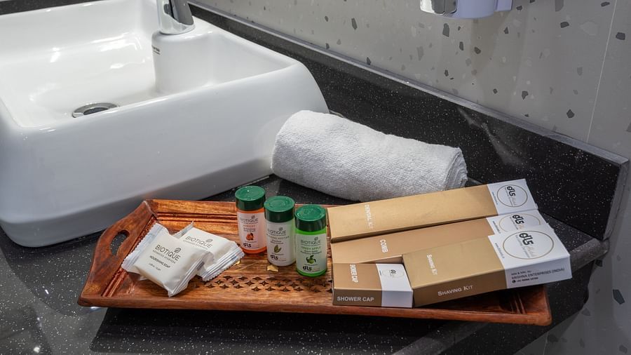  A bathroom countertop with a modern square basin and a wooden tray containing toiletries including small bottles, soap bars, and boxed items; a neatly rolled white towel and a wall-mounted dispenser are also visible - DLS Nature Trinket Resort, Dalhousie.