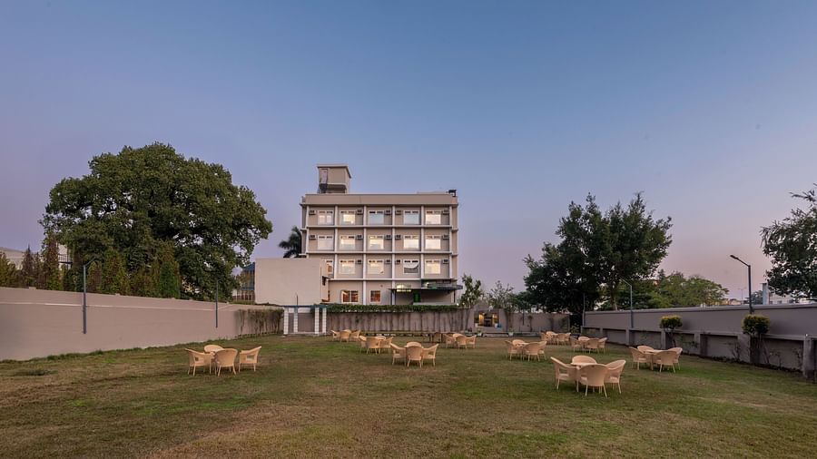 Lawn area of DLS Park Grace, Haridwar featuring seating area featuring tables and chairs, luscious green trees, and a tall building.