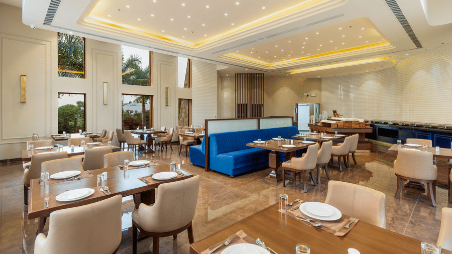 An indoor dining area at EsthereaRaj Leela, Ranakpur, with wooden tables, tan chairs, blue booth seating, and recessed ceiling lighting.
