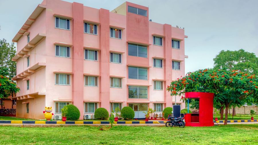 A vibrant exterior shot of a pink hotel building with greenery and a red security booth in the foreground at Greens Inn - Inviting Hotel in Hyderabad.