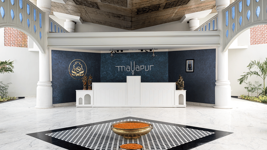 A reception lobby at Hotel Sonar Bangla, Mayapur, featuring a white counter, brass bowls on patterned floor tiles, and architectural arches.