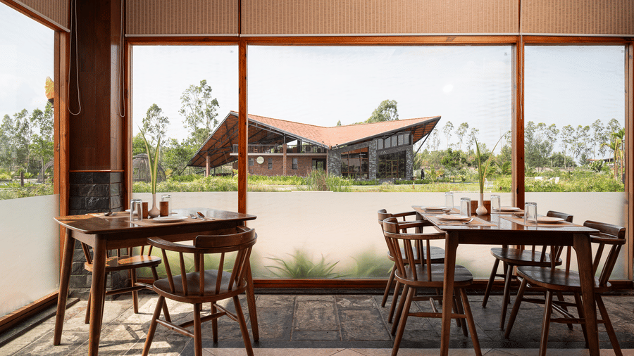 2 wooden dining tables set with glassware and cutlery positioned next to a large window showing an external building with a slanted red roof at Hotel Sonar Bangla, Sundarban. 