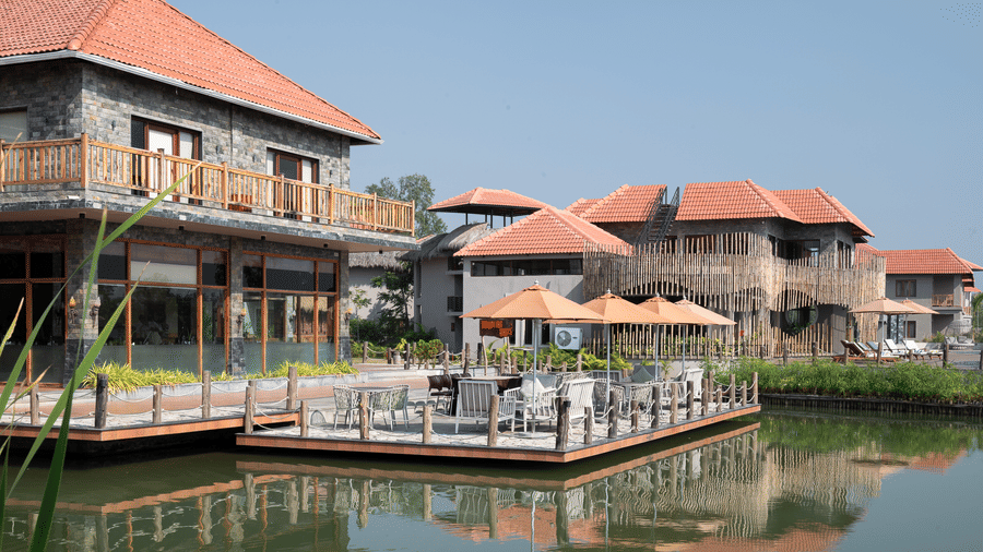 2-storey stone building with a red tiled roof and wooden balconies situated next to a pond with a floating wooden deck and patio seating at Hotel Sonar Bangla, Sundarban.