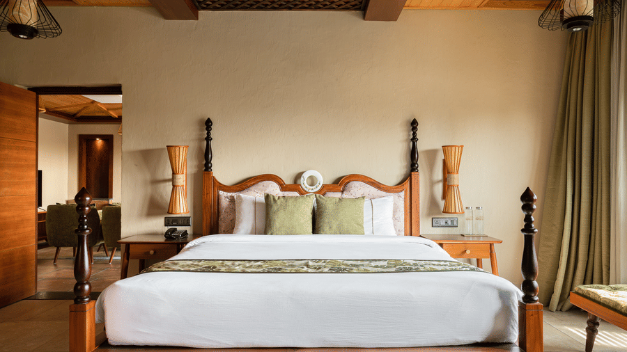 A room at Hotel Sonar Bangla, Sundarban with 4-poster king bed, wooden coffered ceiling, lantern wall lights, and adjoining living area visible through doorway.