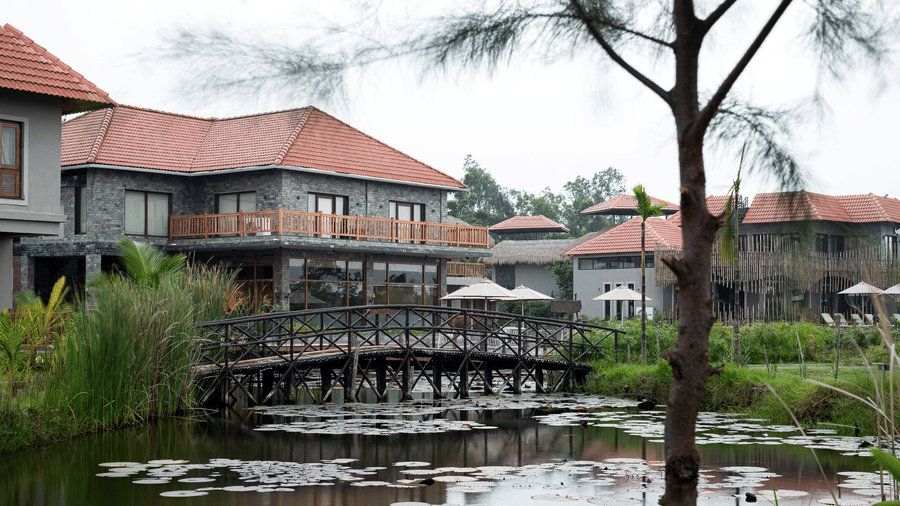 Hotel Sonar Bangla, Sundarban exterior view with stone-clad buildings, terracotta roofs, lily pond, wooden footbridge, parasols, and surrounding greenery.