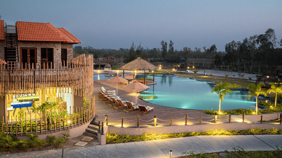 Evening view of the outdoor swimming pool at Hotel Sonar Bangla, Sundarban with thatched gazebo, sun loungers, trees, and a tiled deck lit by lamps.
