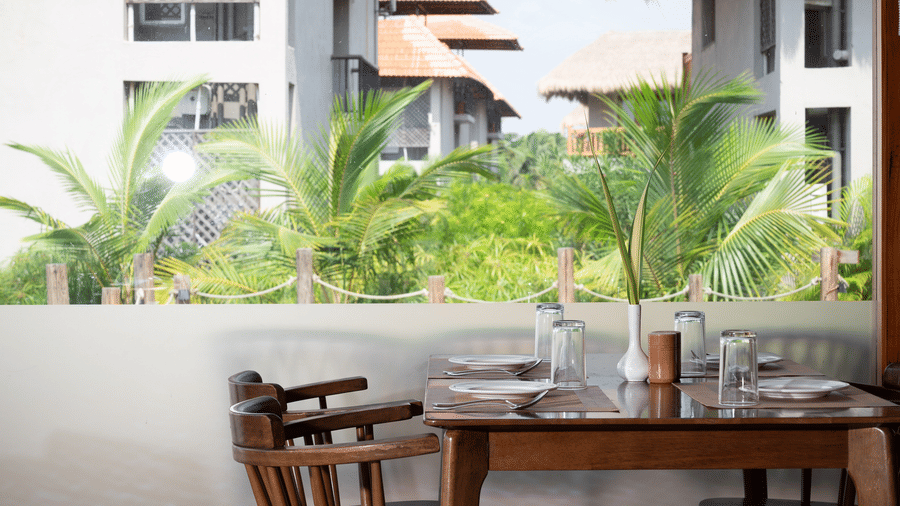 Wooden dining table for 2 set against a low wall with green trees and hotel buildings in the background at Hotel Sonar Bangla, Sundarban.