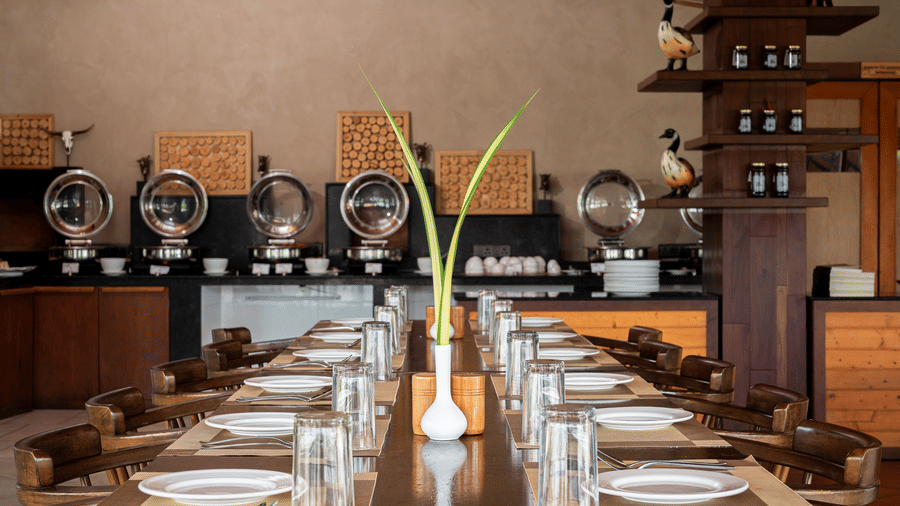 Long wooden dining table set for a meal with a buffet counter featuring silver chafing dishes in the background at Hotel Sonar Bangla, Sundarban.