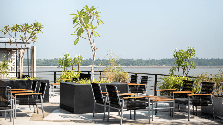 Wide view of the open-air rooftop seating area with multiple groups of modern outdoor tables and chairs, overlooking the water in the distance at Hotel Sonar Bangla, Taki.