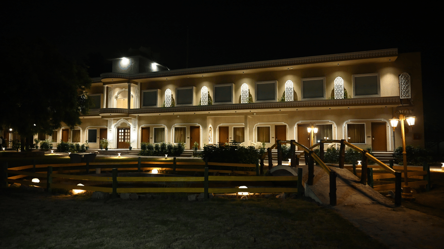 The two-story white building of Kadamb Kuteer at night, showing symmetrical windows, a green lawn in the foreground, and glowing exterior lights.