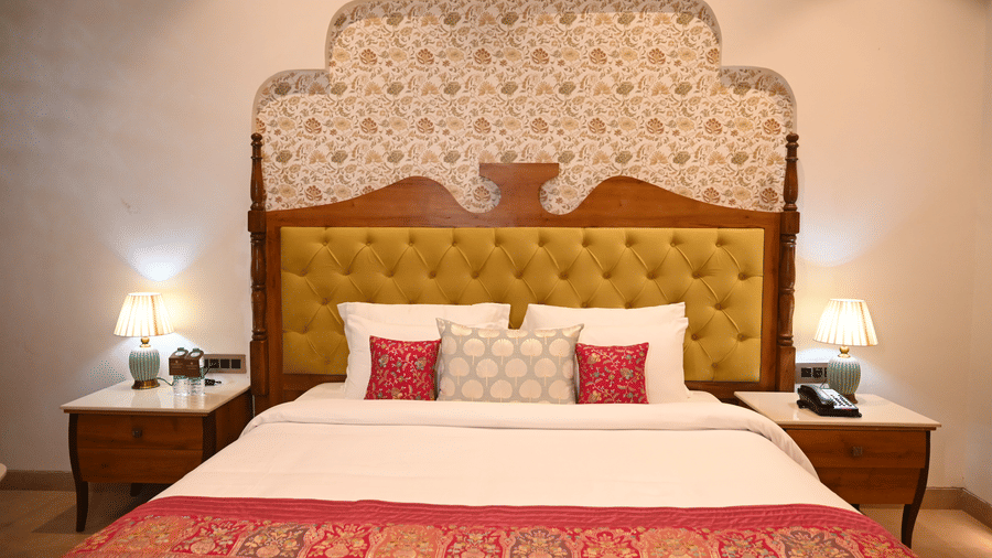 A centered view of a bed at Kadamb Kuteer with a large decorative headboard, white bedding, pink accent pillows, and wall-mounted reading lamps.