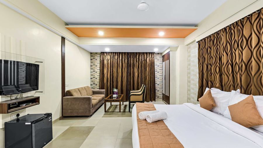 A well-lit suite in Krushnai Resort featuring a double bed with blue runner, a wall mounted TV, docking station, couch and a lounge chair.