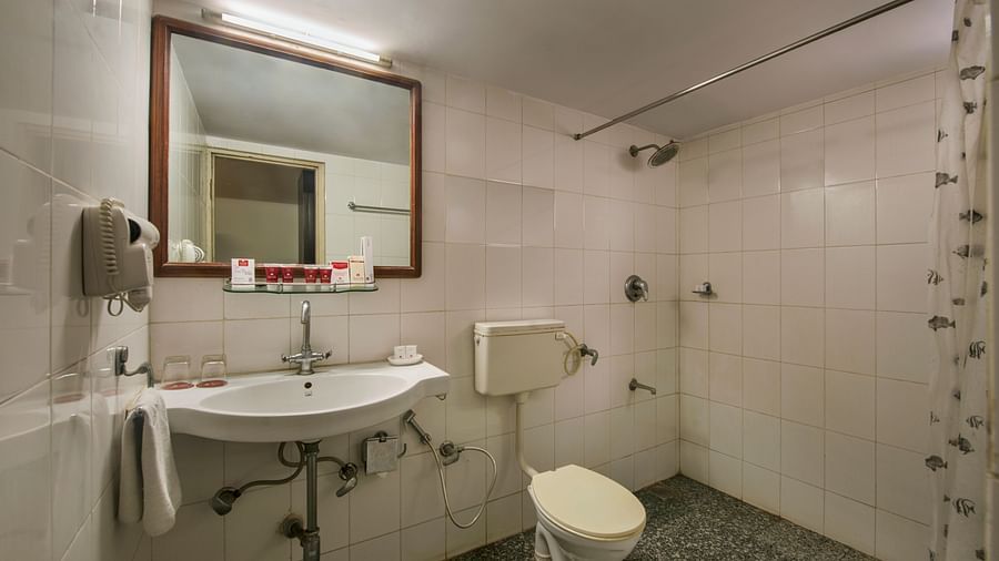 A small bathroom with a white sink, toilet, a mirror with a wooden frame, and a shower area with a patterned curtain - Lords Eco Inn Dahej