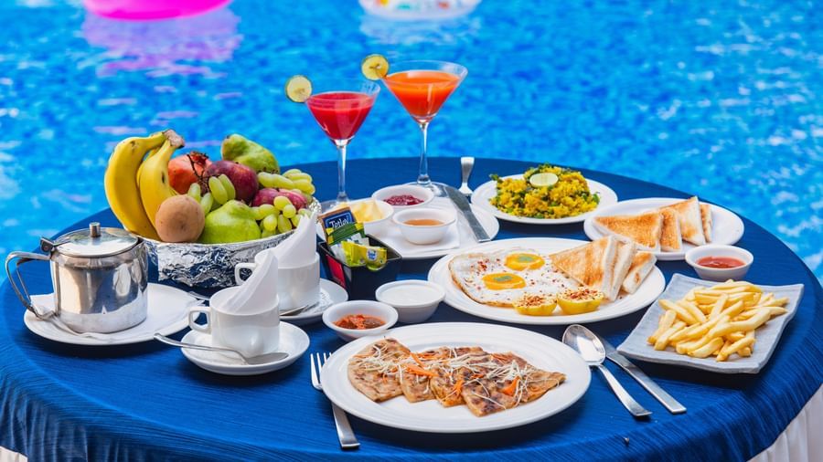A round table besides the pool with several food items displayed on it, at Mayur Retreat.