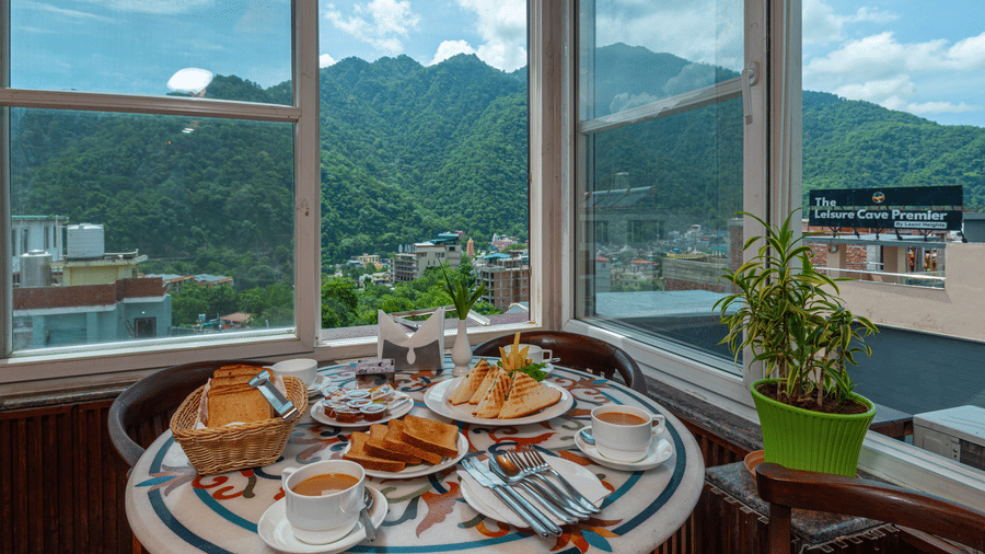 A cafe table is set for breakfast, with plates of toast, grilled sandwiches, jam, and coffee. The setting is bright with natural light from large windows, offering a view of a green, forested mountain and a townscape below. This is a shot from the cafe at 