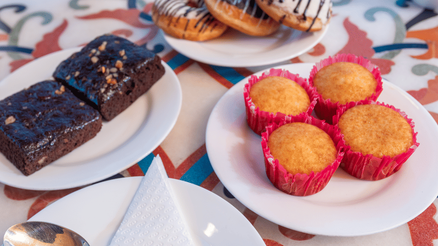 A close-up shot of a table with plates of baked goods, including chocolate donuts with white drizzle, chocolate brownies with nuts, and four cupcakes in pink paper liners. This is a food shot from the cafe at DLS Hotels On The Ganges.