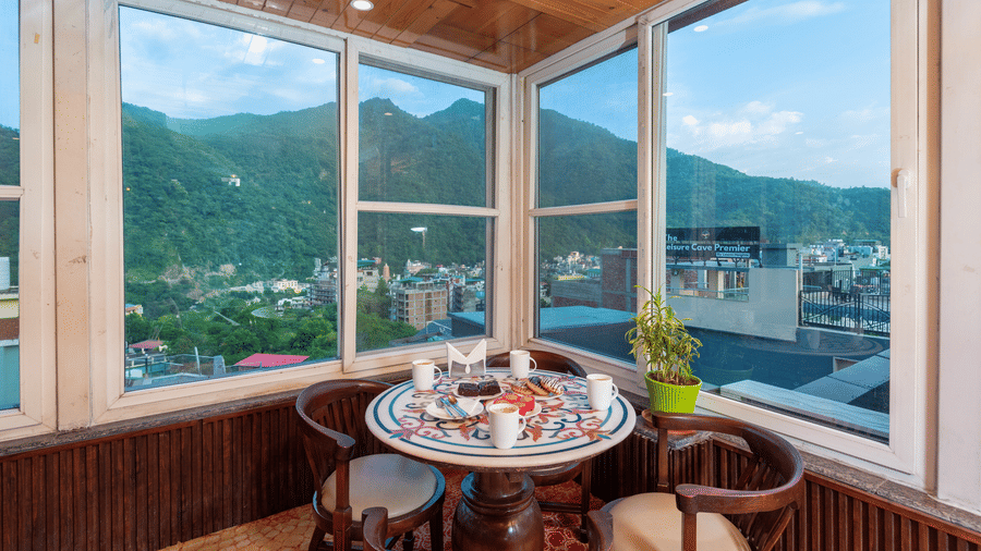 A cafe dining area with a round table set for a meal, including drinks, a cake, and pastries. The room has large windows offering a panoramic view of lush green hills and the town below, with a wooden-paneled ceiling and walls. This is a dining area at Reg