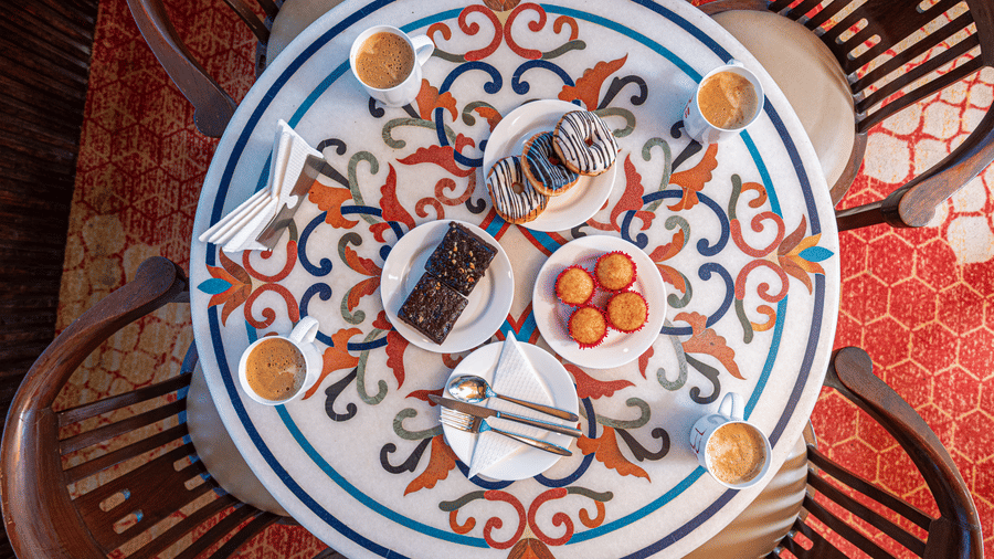 An overhead shot of a circular table set with a variety of pastries, brownies, and four cups of coffee. The table has a colorful, intricate floral and geometric pattern, with four chairs arranged around it. This is a food shot from the cafe at DLS Hotels O
