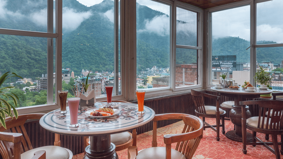 A cafe with a vibrant, red patterned carpet and wooden furniture. The room has large windows that look out onto misty green mountains and the city below. Tables are set with various drinks and food, including a fruit platter and sandwiches. This is the caf