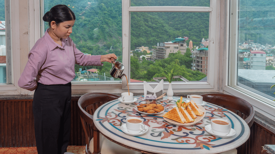 A woman in a pink shirt and black pants is pouring tea from a silver pot into a white cup at a cafe table. The table is set with cookies and a plate of grilled sandwiches. The background features a large window with a view of misty, green hills. This is a 