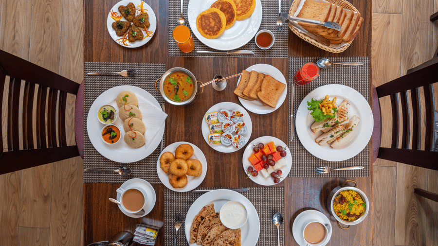 An overhead shot of a dining table laden with a variety of breakfast items, including pancakes, toast in a basket, grilled sandwiches, a platter with idli and chutneys, donuts, parathas with curd, and various juices and coffee. This is a breakfast spread f