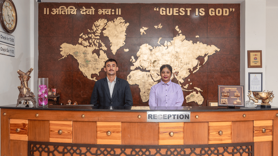Two hotel staff members, a man and a woman, stand behind a wooden reception desk. Behind them is a large wall with a world map and the phrase Guest Is God in English and Hindi. This is the reception area at DLS Hotels On The Ganges.