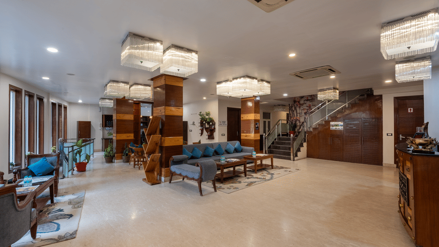 A wide shot of a modern hotel lobby and seating area. The space features a grey sofa with blue pillows, two low coffee tables, and a long bench. Wooden columns with recessed lighting add warmth to the space, and a staircase is visible in the background. Th