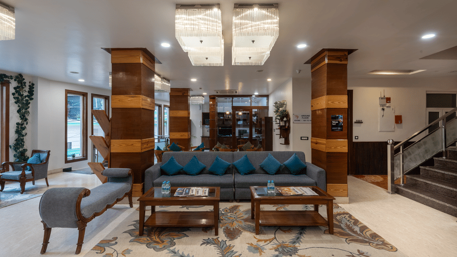  A wide shot of a modern hotel lobby with a seating area. There is a large grey sofa with blue pillows and two wooden coffee tables on a patterned rug. The space is well-lit with elegant, modern chandeliers and features wooden accents on columns and walls.