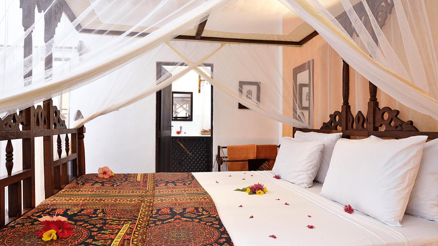Side view of a bed with a mosquito net, decorative bedding and flower petals placed on it. A doorway leading to bathroom can be seen in the background - Shooting Star Boutique Hotel.