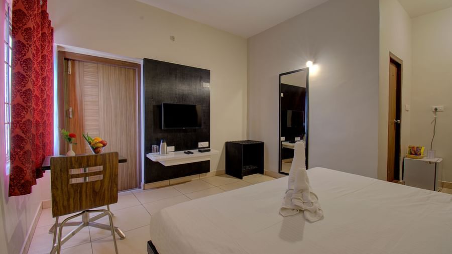 A bedroom with a bed, chair, TV unit, curtained windows, mirror and wooden door | Superior Room | woodstock resorts, Coorg