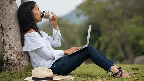 An image showing a woman drinking a cup of coffee, while sitting on the grass with her back on a tree, with a fedora hat and a laptop with her, and bushy landscapes in the backdrop