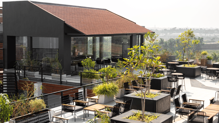 Wide view of the Hotel Sonar Bangla, Taki, rooftop area, featuring an enclosed structure with a sloped roof, surrounded by numerous outdoor tables, chairs, and planter boxes.