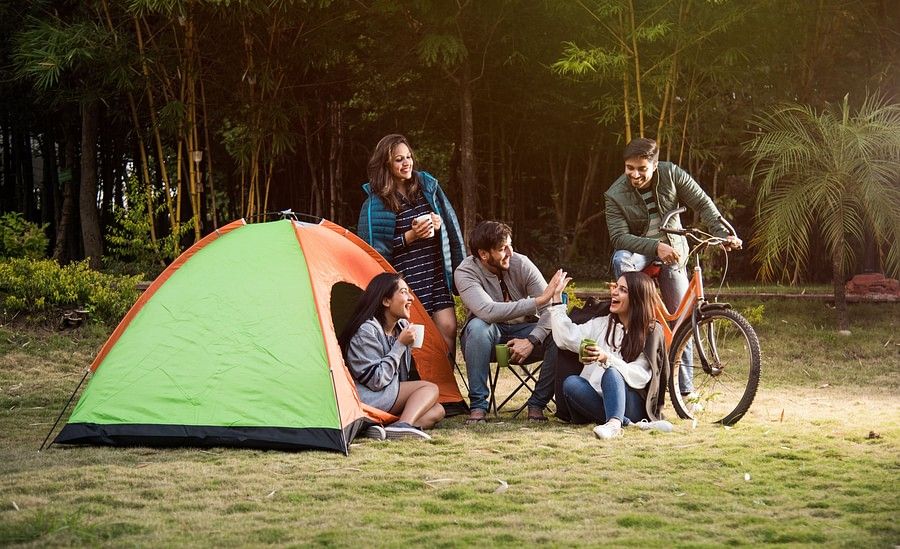  A group of six people, including adults and teenagers, are gathered around a campsite with a green and orange tent. They are enjoying a meal and drinks together, with bicycles parked nearby.
