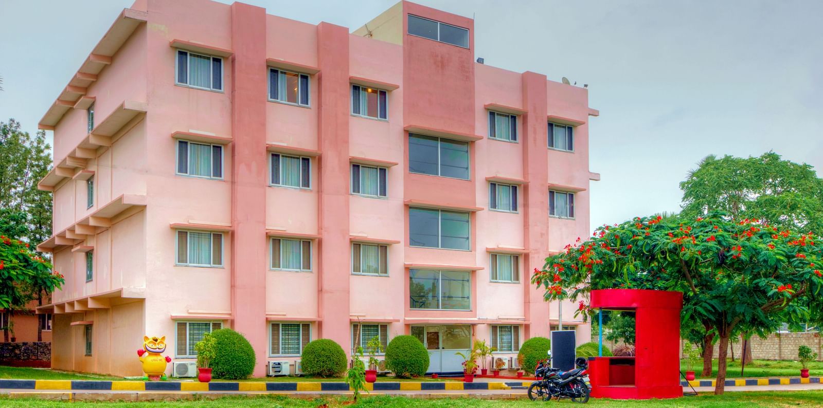 A vibrant exterior shot of a pink hotel building with greenery and a red security booth in the foreground at Greens Inn - Inviting Hotel in Hyderabad.