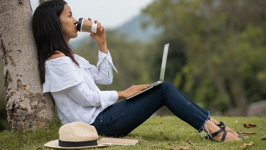 An image showing a woman drinking a cup of coffee, while sitting on the grass with her back on a tree, with a fedora hat and a laptop with her, and bushy landscapes in the backdrop