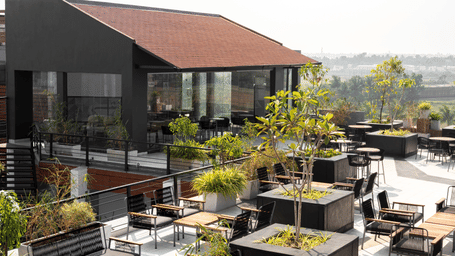 Wide view of the Hotel Sonar Bangla, Taki, rooftop area, featuring an enclosed structure with a sloped roof, surrounded by numerous outdoor tables, chairs, and planter boxes.
