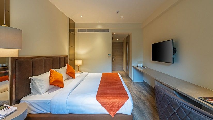 A plush bed room with pillows, runner, a TV mounted on the wall in front of it and a hallway with a door at Hotel Saket 27, New Delhi