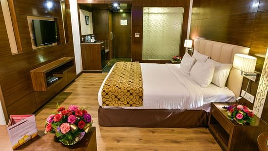 A well-appointed hotel room with wooden accents features a bed with multiple pillows, bedside tables with lamps and flowers, a television, and a glimpse into an adjoining area - Renest Jaipur
