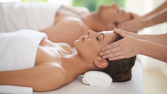 A couple laying on two white massage beds, wearing white towels and facing upwards, while their face is being massaged by two practitioners.