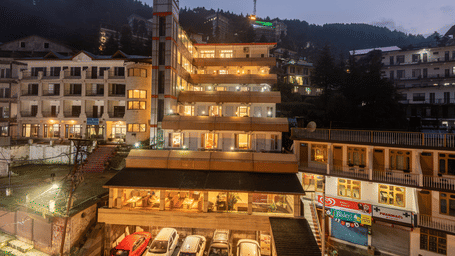 Exterior facade view of DLS Spring Valley Resort, Dharamshala, featuring bright lighting and a car parking area below.