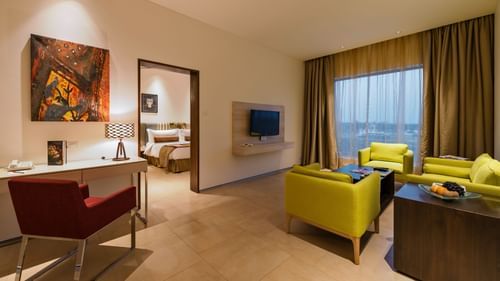An image of the separate living room of the Suite with sofa set, centre table, work desk, chair, flat screen TV, and an expansive window with curtains - Pride Elite Phaltan